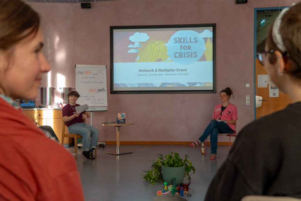 There is a screen with a presentation projected on it. There is a logo of the project skills for crisis on it and the name of the event "network and multiplier event". There are two people sitting under the screen on the chairs. There are also two people closer to the camera, one person is looking at another one.