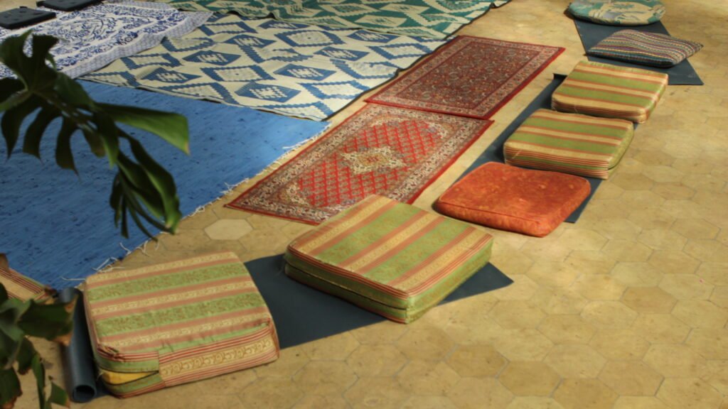 A variety of cushions and carpets that are place in a circle are one the floor. There is a glimpse of light shining on them through the windows of the chapel. 