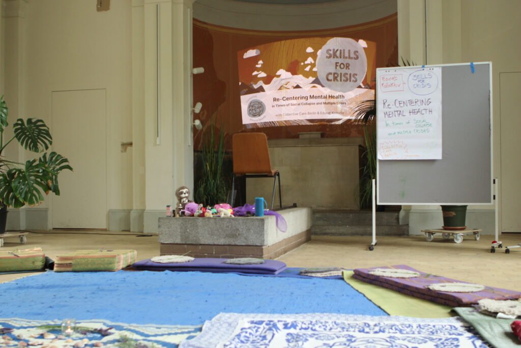 The picture shows the set-up of the room where the workshop took place – an old chapel in Berlin that was never baptized and is now used for community events. On the ground, there are carpets and pillows for people to sit. There is a pinboard with a flipchart with the title of the workshop. On a small platform, there is a variety of stimming tools and teddy bears for comfort and stimming. On the back of the room, where the altar used to be, the presentation of the project in it´s visual design is projected. 