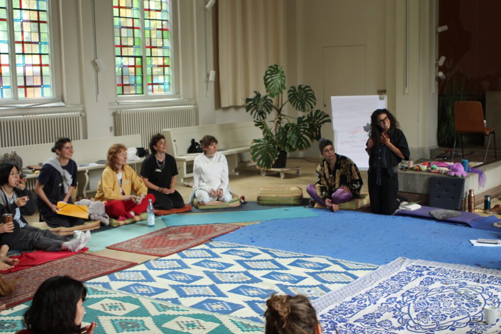There is a group of people sitting in a circle on cushions on the floor. Part of them are participants of the workshop, parts are the facilitators. From what we know from the workshop, many but not all of them were white women and queers from different countries. They all look rather curiously towards something or someone outside of the frame, maybe someone share a story.