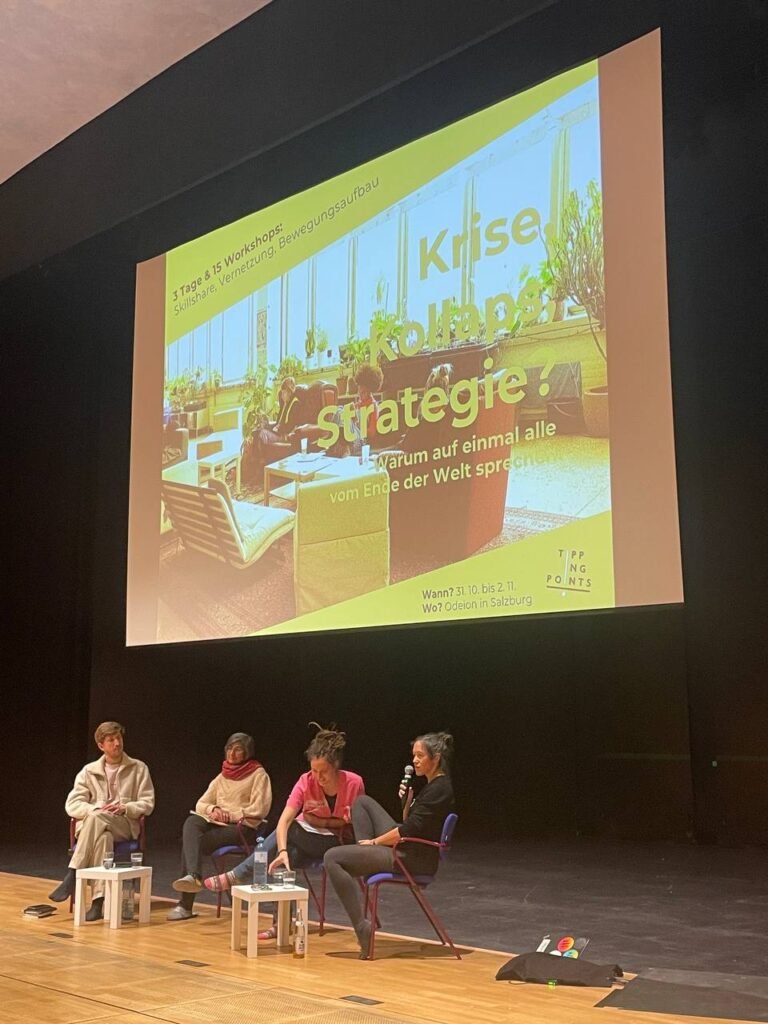 There is a wooden stage and four people are sitting on the chairs on ir engaged in a conversation. There is a big screen behind them with a presentation on it.