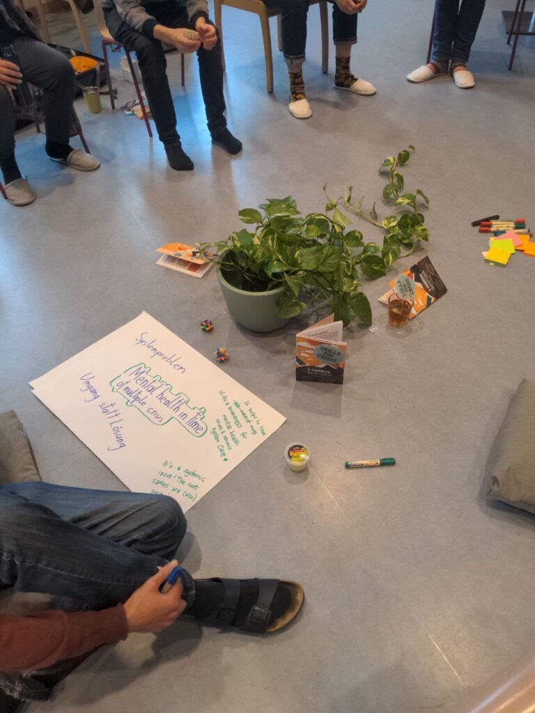 In the picture it is visible , that people are sitting in the circle on the chairs. One person is sitting on the floor, only legs are visible. In the middle on the floor there is a pot with a green flower. There are some handbook around it and some markers with posteds. There is a big flipchart paper with notes written on it about the mental health workshop.