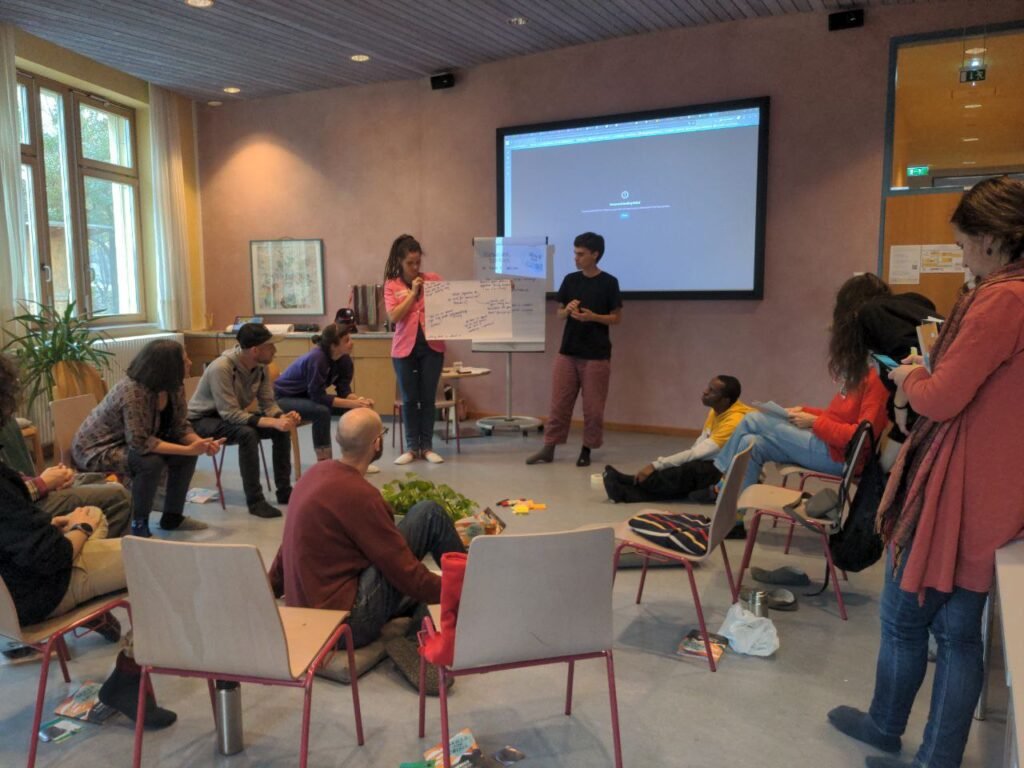 In the picture there  are some people sitting in a circle on the chairs and two people are standing. They hold a white flipchart paper with notes. They present the result of smaller group work. Behind them there is a screen with a black presentation screen.