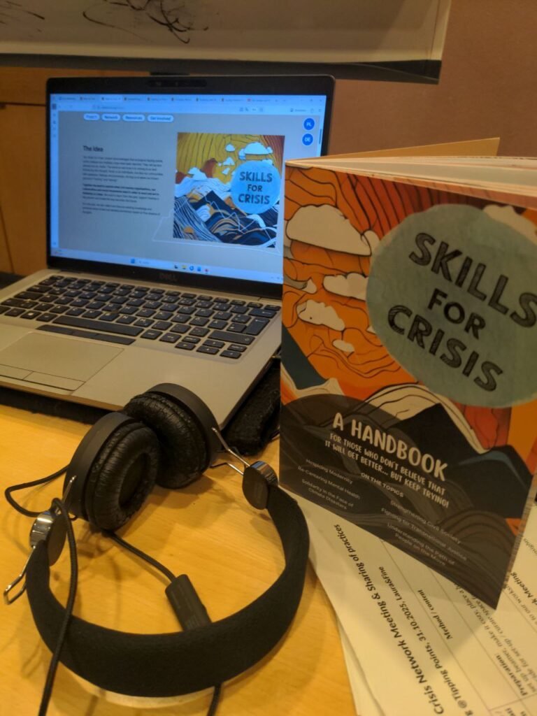 In the picture there is a laptop a bit in the back with a website skills for crisis opened on it. More in front there is a handbook standing on a few white sheets of paper. Ther eare also black headphones lying on the table.