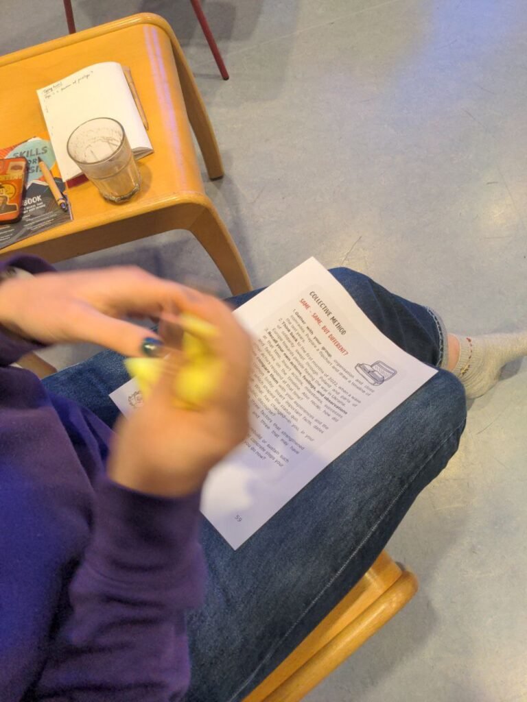 There is a picture of the person holding a sheet of paper on their legs. On the paper there is a collective method description and invitation to practice it. The person is holding a yellow stimming toy in their hands. There is also a chair visible at the top of the picture with a handbook, a notebook and a glass lying on it.